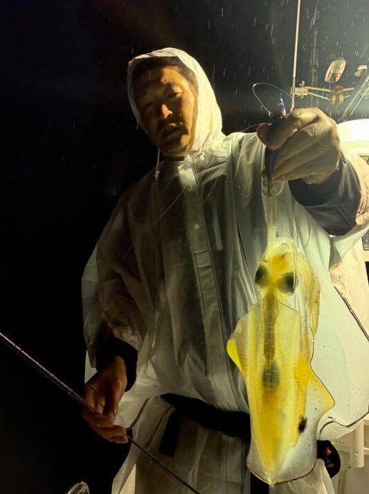 鳥取のティップランでアオリイカを釣り上げたお客様。白いレインウェアを着用し、雨が降る中での貴重な一杯を手にポーズ。