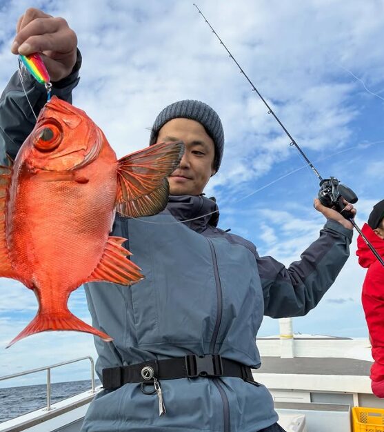 鳥取の遊漁船でのジギング釣果。灰色のウェアにニット帽をかぶった釣り人が、虹色のメタルジグで釣り上げた鮮やかな赤色の根魚（アカムツ）を、ロッドと共に手にしている。