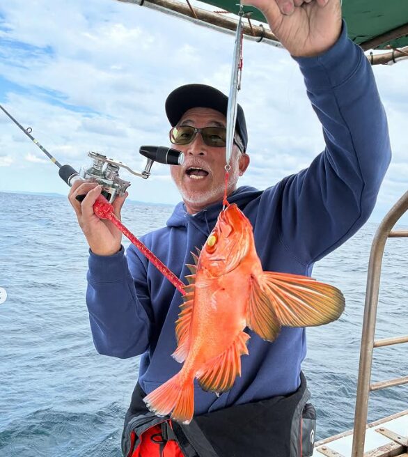 鳥取の遊漁船でのジギング釣果。青いパーカーにサングラス姿の釣り人が、シルバーのメタルジグで釣り上げた鮮やかな赤色の根魚を、ロッドと共に嬉しそうに掲げている。