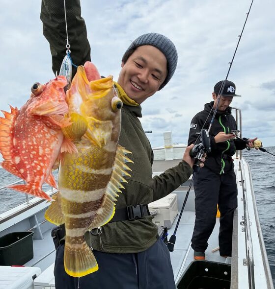 鳥取の遊漁船でのジギング釣果。ニット帽をかぶった釣り人が、1つのジグで同時に釣り上げたとみられる2匹の根魚（カサゴとアオハタ）を、ロッドと共に持ち笑顔を見せている。