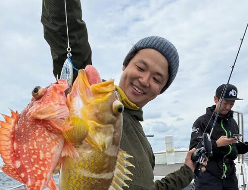 鳥取の遊漁船でのジギング釣果。ニット帽をかぶった釣り人が、1つのジグで同時に釣り上げたとみられる2匹の根魚（カサゴとアオハタ）を、ロッドと共に持ち笑顔を見せている。