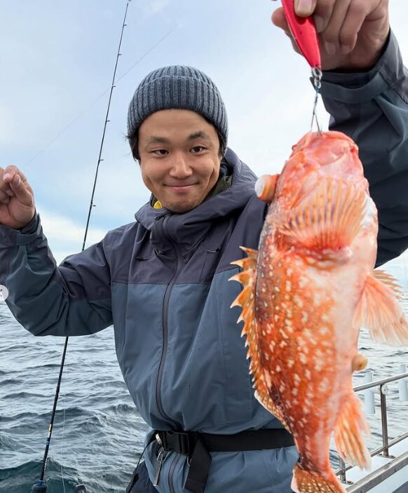 鳥取の遊漁船でのジギング釣果。灰色のウェアとニット帽の釣り人が、赤いメタルジグで釣り上げた根魚（カサゴ）を手に持ち微笑んでいる。