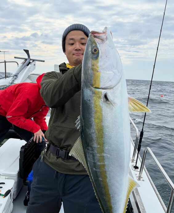 鳥取の遊漁船でのジギング釣果。ニット帽をかぶった釣り人が、釣り上げた立派なマルゴ（ブリの若魚）を両手で掲げている。