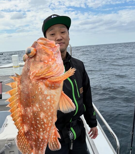 鳥取の遊漁船でのジギング釣果。黒いウェアとキャップ姿の釣り人が、釣り上げた良型（りょうがた）の根魚（カサゴ）を片手で誇らしげに掲げている。