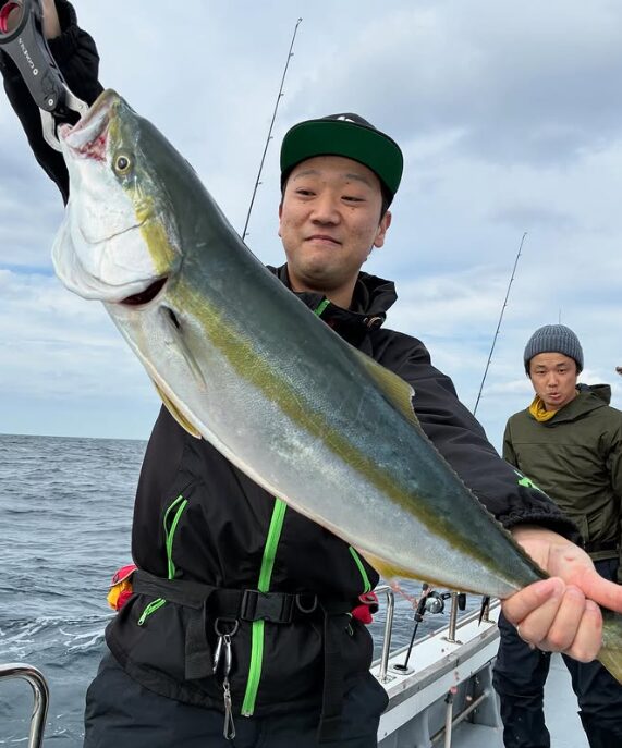 鳥取の遊漁船でのジギING釣果。黒いウェアとキャップ姿の釣り人が、フィッシュグリップを使って釣り上げたマルゴ（ブリの若魚）を笑顔で掲げている。