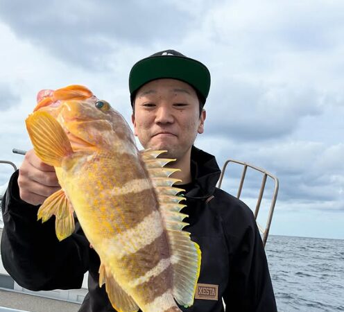鳥取の遊漁船でのジギング釣果。黒いウェアとキャップ姿の釣り人が、釣り上げた鮮やかな黄色い根魚（アオハタ）を顔の横で掲げている。