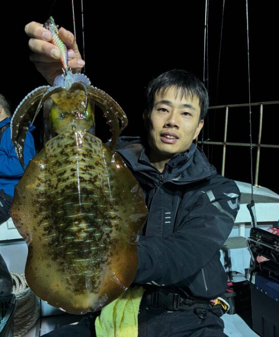 夜の船上で、大きく立派なアオリイカを手に満足そうな釣り人。ティップランでの素晴らしい釣果を収めた様子。