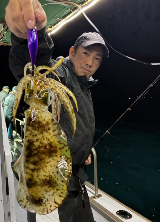 夜の船上で、ティップランで釣り上げたアオリイカを手に持つ釣り人。見事な釣果をカメラに向ける様子。