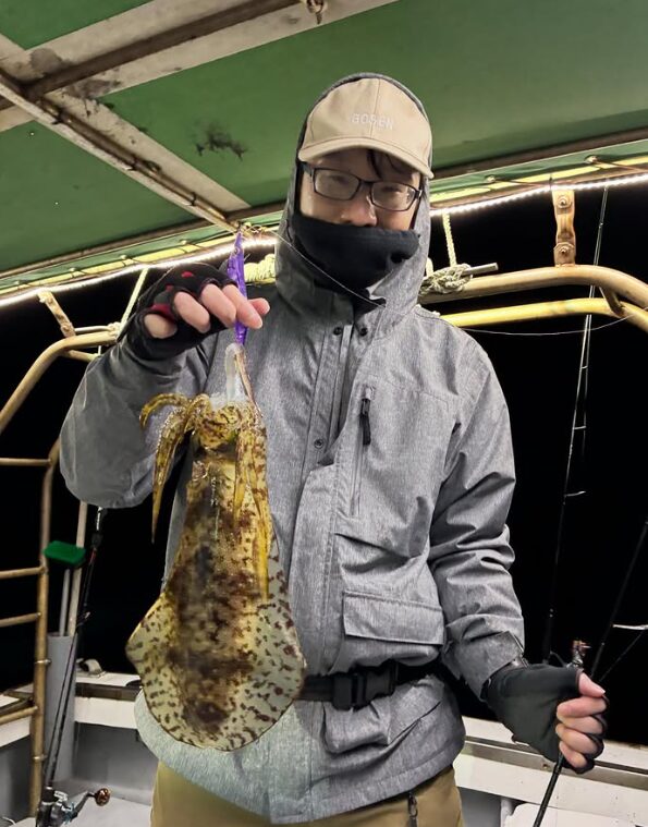 鳥取のアオリイカティップランにて、ベージュのキャップとグレーのジャケットを着用したお客様が、紫系のエギで良型アオリイカをキャッチした様子