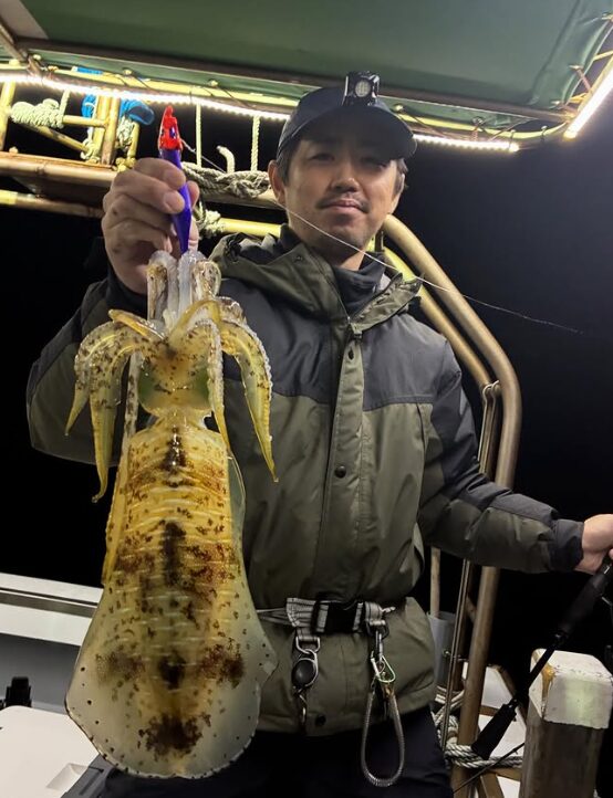 境港遊漁船の夜釣りにて。カーキ色のジャケットを着てヘッドライトを装着した男性が、ティップランで紫のエギを使ってキャッチした立派なアオリイカを見せている様子。