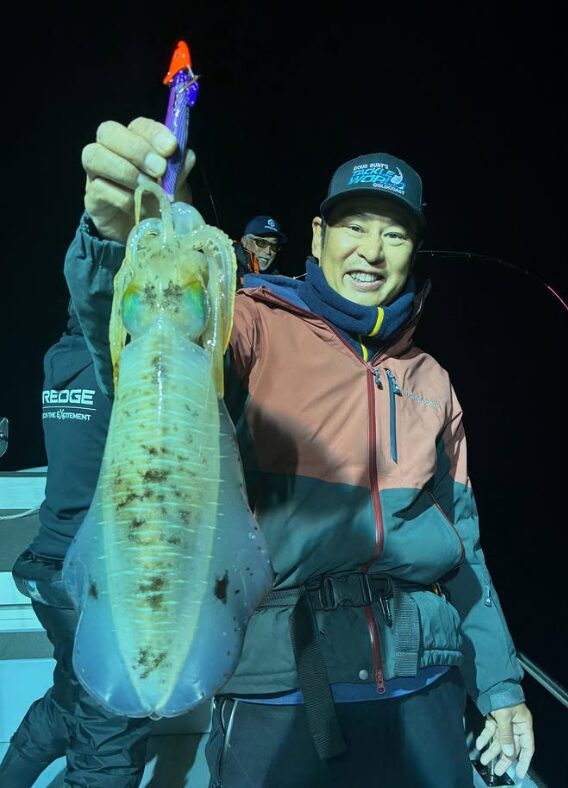 境港遊漁船の夜便にて。赤茶色のジャケットとキャップを着用した男性が、紫色のエギを使ったティップランで釣り上げた良型のアオリイカを手に満面の笑みを見せている様子。背景には他の釣り人も写っている。