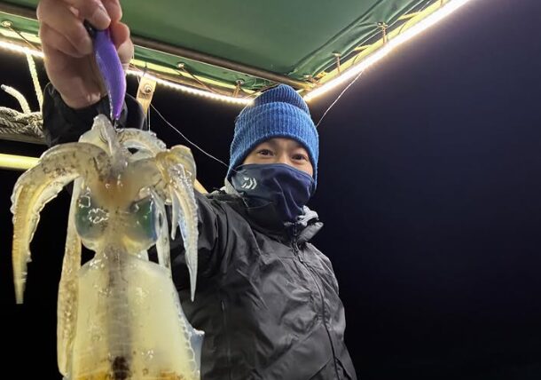 境港遊漁船での夜焚きティップラン釣果。青いニット帽と黒いレインウェアを着用した男性アングラーが、紫系のエギでキャッチした良型のアオリイカを手に持つ様子。