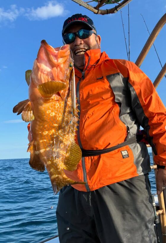 鳥取ジギングでのハイライト。鮮やかなオレンジ色のジャケットを着た男性が、青空の下で特大のアコウ（キジハタ）を抱え、サングラス越しにもわかる満面の笑みを見せている。
