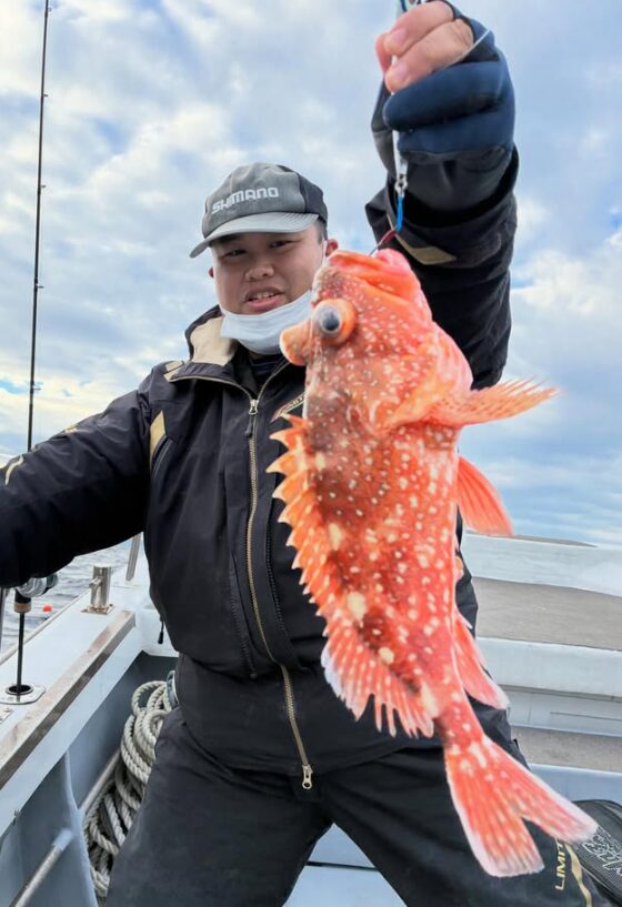 鳥取の根魚ジギング釣行にて。シマノのグレーの帽子と黒にゴールドラインのウェアを着た男性が、ジギングでキャッチした良型のカサゴ（ガシラ）を掲げている写真。