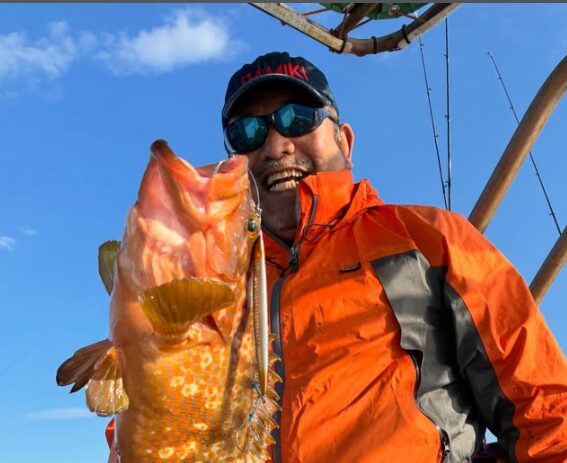 鳥取ジギングでのハイライト。鮮やかなオレンジ色のジャケットを着た男性が、青空の下で特大のアコウ(キジハタ)を抱え、サングラス越しにもわかる満面の笑みを見せている。