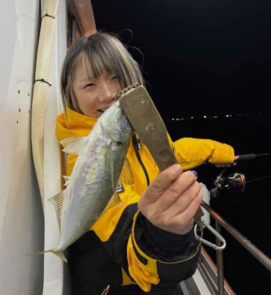 取アジングを楽しむ女性アングラーの釣果。髪に金色のメッシュが入った、黄色いウェアの女性が、フィッシュグリップで掴んだ良型のアジを顔に寄せてニッコリと笑っている。尺アジも期待できる夜釣りでの一枚。