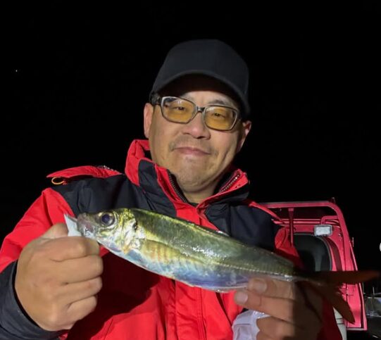 鳥取アジングの釣果写真。赤い防寒ジャケットと黒いキャップ、メガネを着用した男性が、夜の港で釣り上げた尺アジサイズの良型アジを両手で持っている様子。