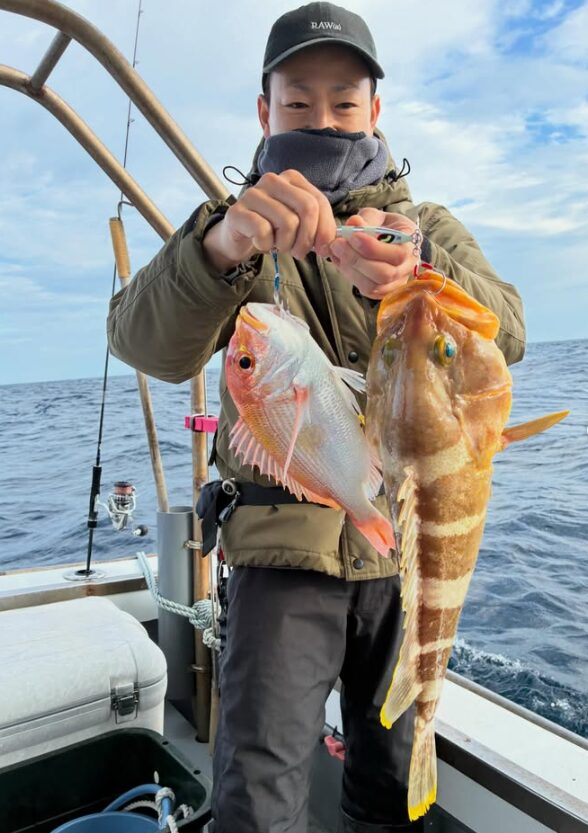 鳥取沖の根魚ジギングにて。黒いキャップとカーキ色のジャケットを着た男性が、メタルジグで釣り上げた良型のアオハタとレンコダイを両手で掲げて持っている様子。