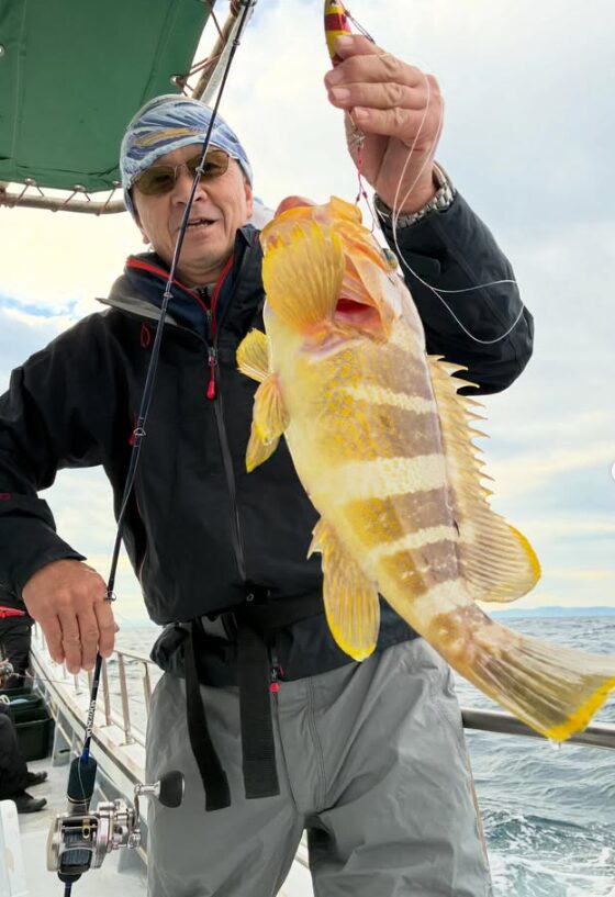 鳥取のジギング船上で、サングラスとバンダナをしたベテラン風の男性が、釣り上げたばかりの鮮やかな良型アオハタを片手で持ち上げている写真。タックルも写っている。
