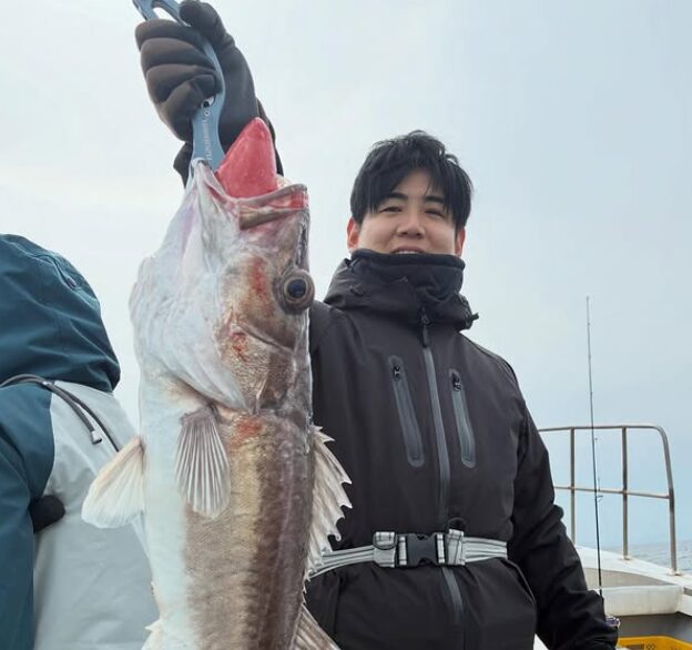境港遊漁船のジギング釣果:黒い防水ジャケットとネックウォーマー、黒いグローブを着用した20〜30代の男性が、青いフィッシュグリップで釣り上げた大きなアラ(根魚)を右手で高く掲げて笑顔を見せている様子