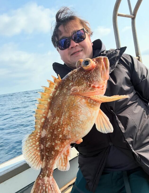 境港の遊漁船にて、サングラスに黒のジャケットを羽織った男性が、スロージギングで釣り上げた良サイズのウッカリカサゴを手に微笑む様子。