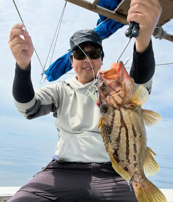 山陰沖のジギングで釣り上げられた珍しい根魚のホウキハタ。黒いキャップとサングラスにグレーのパーカーを着用した男性が、特徴的な縞模様を持つ良型のホウキハタをフィッシュグリップで高く掲げて笑顔を見せている船上の写真。