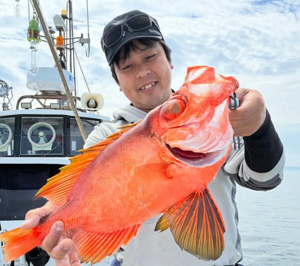 山陰沖のジギング釣果。遊漁船のキャビンを背景に、サングラスを黒いキャップの上に乗せたグレーのパーカー姿の男性が、釣り上げた色鮮やかで大きな赤い根魚を両手でしっかりと横に構え、嬉しそうに微笑んでいる写真。